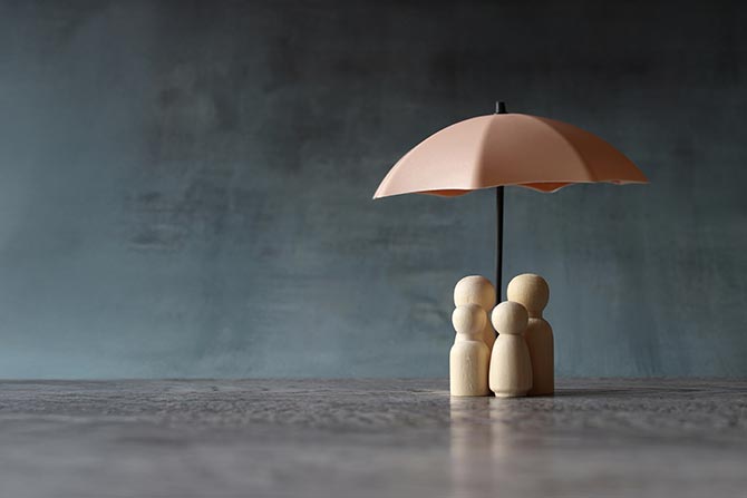 Finding Common Ground on Deposit Insurance Reform; Four wooden figurines huddle under a small pink umbrella on a textured surface. The background is a moody gray, conveying protection and unity.