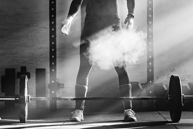 It’s Time To Hit the Weights; Person stands over a barbell in a dimly lit gym, surrounded by chalk dust and sun rays. The scene conveys determination and focus.