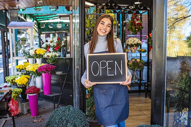 From the Top: It’s a Wonderful (Community Banking) Life; A smiling woman in an apron stands at a flower shop entrance, holding an "OPEN" sign. Vibrant flowers surround her, evoking a welcoming atmosphere.