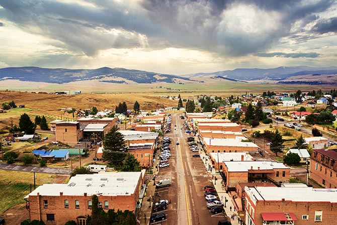 Main Street Capital Access (“Main Street”) Act; Aerial view of a small town with a central road lined by buildings and parked cars. Surrounding fields and distant mountains under a cloudy sky. Calm and rustic atmosphere.