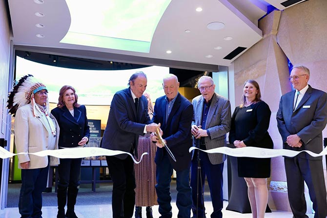 Montana Heritage Center Opens to Public After 20-Year Journey; A group of seven people in formal attire stand together in a brightly lit room for a ribbon-cutting ceremony, smiling and holding scissors. The setting appears modern and celebratory.